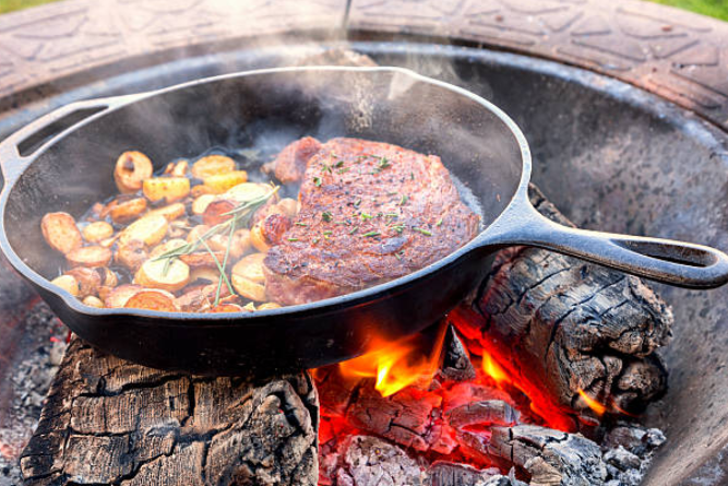 Steak and small potatoes being cooked in a cast iron skillet over an open flame fire.
